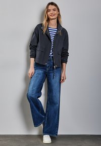 Marineblauwe lichte bomberjack met ritssluiting, gecombineerd met brede blauwe jeans en witte sneakers. Gestreept shirt zichtbaar eronder.