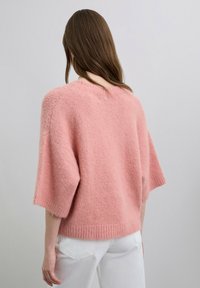 Suéter de punto rosa claro con mangas cortas, con dobladillo acanalado y textura suave. Se puede ver un pequeño detalle de botón en la parte trasera.