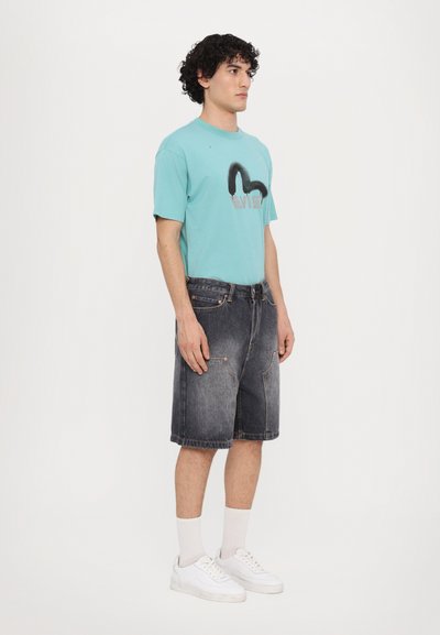 T-shirt grafica turchese con design nero e rosso, abbinata a pantaloni shorts di jeans scuri e sneakers bianche con calzini a coste.