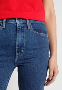 Modré džínové kalhoty s vysokým pasem, hladkou texturou, klasickým designem s pěti kapsami a zapínáním na knoflík v pase.