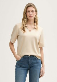 Camisa polo beige con cuello en V, mangas cortas y textura suave, combinada con jeans de mezclilla azules. Diseño sencillo sin patrones visibles.