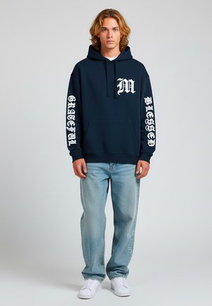 Felpa blu marina con testo grafico bianco, tasca frontale e cappuccio con cordino, abbinata a jeans azzurri e sneaker bianche.
