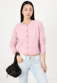 Ανοιχτό ροζ πουλόβερ από μάλλινο ύφασμα με στρογγυλή λαιμόκοψη και κλείσιμο με έξι κουμπιά, συνδυασμένο με ανοιχτό μπλε τζιν και μια μαύρη τσάντα με κορδόνι και λουλουδάτο μοτίβο.