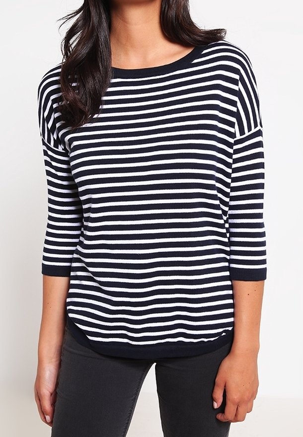 Femme portant une chemise à rayures horizontales bleu marine et blanche à manches trois-quarts ainsi qu'un pantalon noir, debout devant un fond uni.