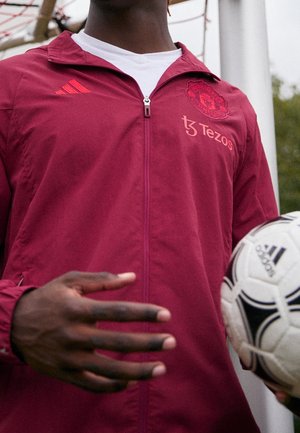 Person wearing maroon Manchester United jacket holding a white Adidas soccer ball near a goalpost outdoors.