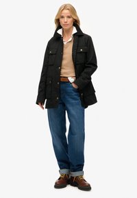 Chaqueta de algodón negra con un cuello alto, múltiples bolsillos frontales y botones de latón, combinada con un suéter beige, jeans azules y botas marrones.