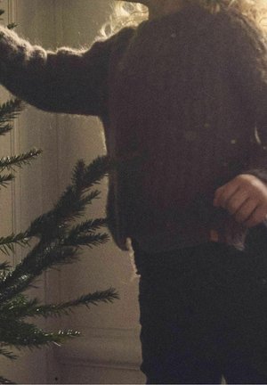 Enfant en pull sombre tendant la main pour décorer un sapin de Noël non décoré à l'intérieur, avec une lumière douce en arrière-plan.