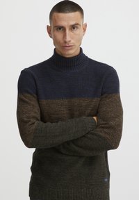 Maglione a collo alto lavorato a maglia in blu scuro, marrone e verde. Texturizzato, con polsini e orlo a coste. Presenta un piccolo logo sul lato.