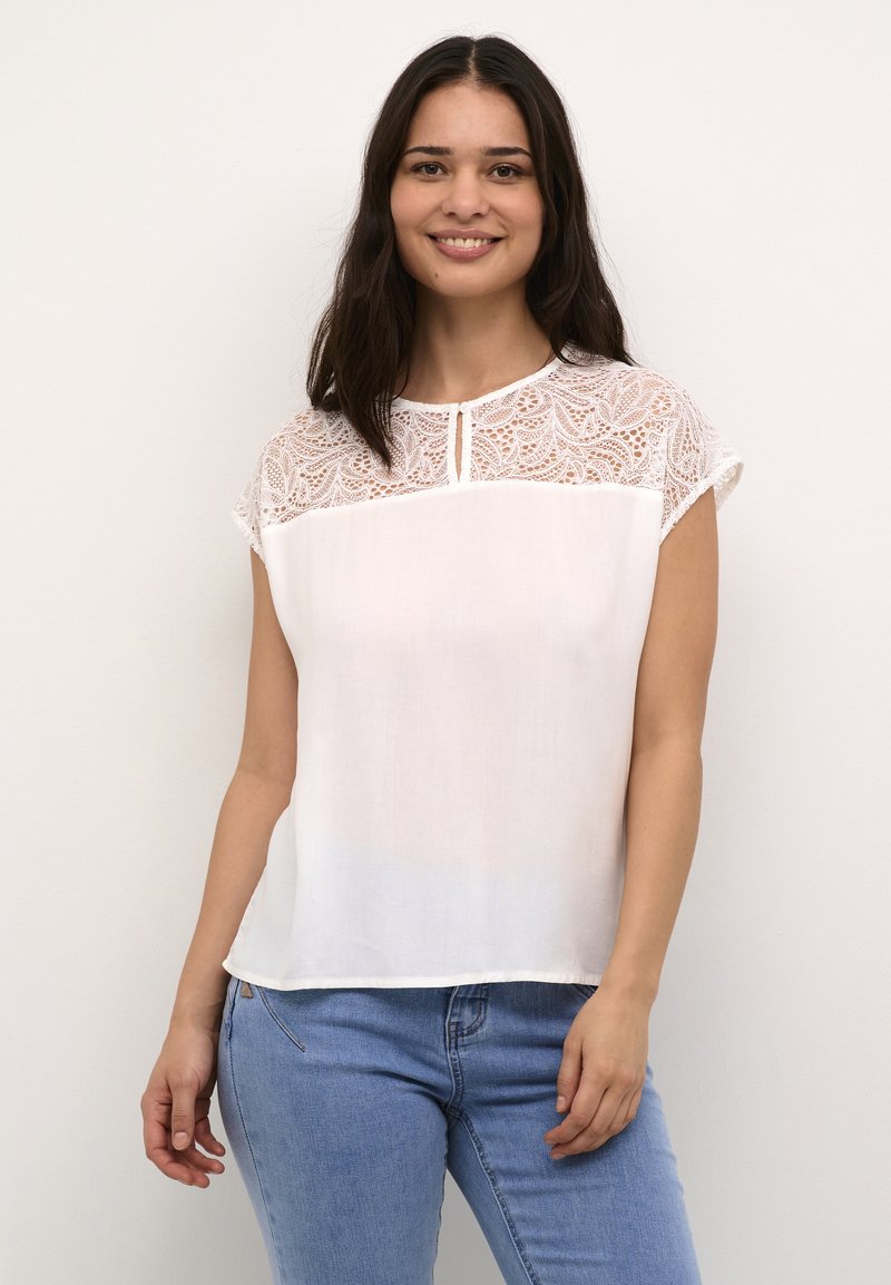 Blusa bianca con yoke in pizzo, maniche corte e un piccolo dettaglio a forma di chiave sullo scollo. Abbinata a jeans blu.