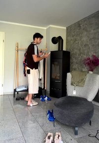 Homme debout près d'un porte-vêtements tenant un téléphone, avec des chaussures au sol, une cuisinière et une chaise grise avec des vêtements pliés dans une pièce moderne.
