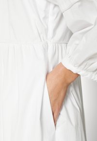 Main insérée dans la poche latérale d'un vêtement blanc à manches longues avec taille froncée et manches à revers.