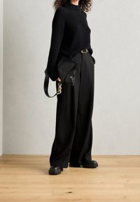 Pull noir à côtes avec col montant, pantalon noir ample, et une pochette en cuir noir avec une chaîne dorée. Chaussures à plateforme noires épaisses.