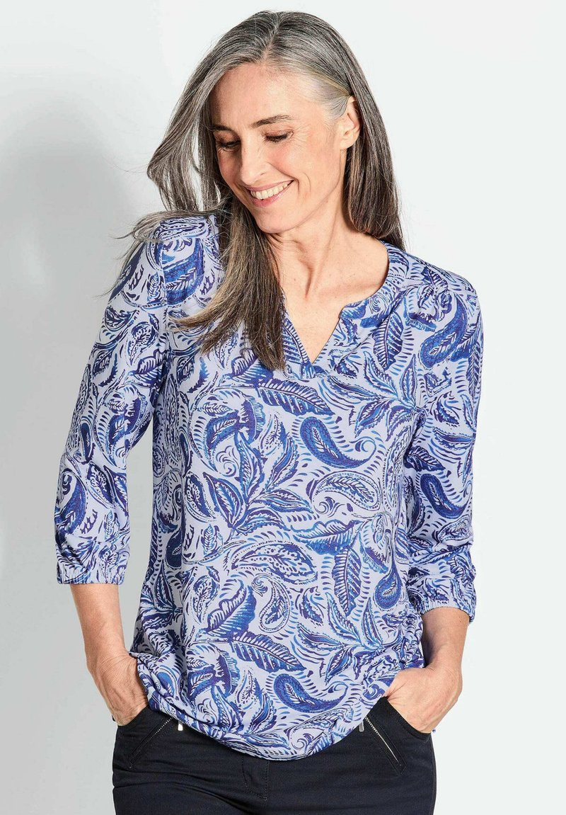 Langärmlige Bluse mit V-Ausschnitt in Hellblau, mit einem Paisleymuster in Dunkelblau und Aquamarin. Merkmale sind aufgerollte Ärmel und eine lockere Passform.