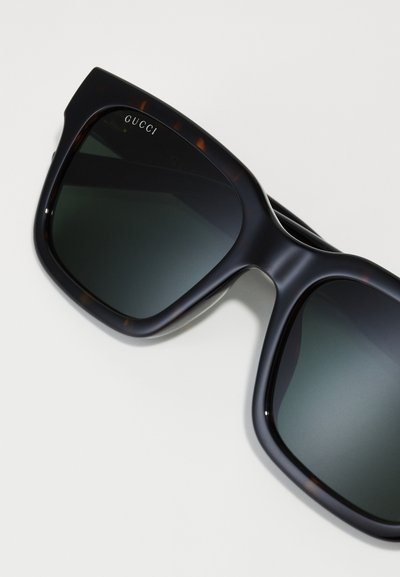 Mustade mustritega ülev suurte prillide klaasid tumehallid ja säravad. Vasakus ülanurgas on hõbedane "GUCCI" logo.