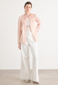 Camicia button-up rosa chiaro con dettagli arricciati, abbinata a pantaloni a gamba larga bianchi e tacchi di colore neutro su uno sfondo semplice.
