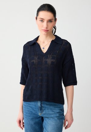 Vrouw met donker haar die een marineblauwe gebreide korte mouw trui en blauwe spijkerbroek draagt, geaccentueerd met een gouden ketting met een blauw hangertje.