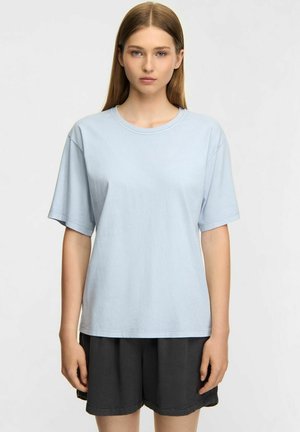 Young woman with straight light brown hair wearing a light blue short-sleeve t-shirt and black shorts, standing against a plain background.