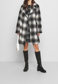 Soprabito a quadri in bianco e nero con design doppiopetto, ampi revers e tasche laterali, indossato sopra un vestito a quadretti e stivali al ginocchio.