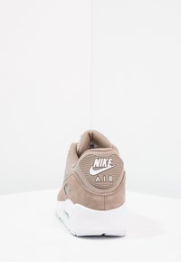 Bežové tenisky Nike Air z broušené kůže s polstrovaným límcem, bílou podrážkou a logem na patě. Texturovaný svršek a hladké akcenty.