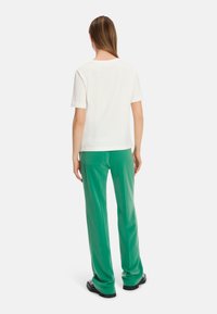 Weißes Baumwoll-T-Shirt mit Rundhalsausschnitt, kombiniert mit grünen, weit geschnittenen Hosen mit einer Gesäßtasche und glatter Textur, gestylt mit schwarzen Schuhen.