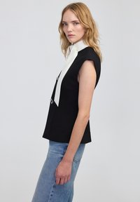 Czarny top bez rękawów z białym kołnierzem i srebrnymi elementami, o dopasowanym kroju i gładkiej teksturze. Noszony z jasnoniebieskimi dżinsami.