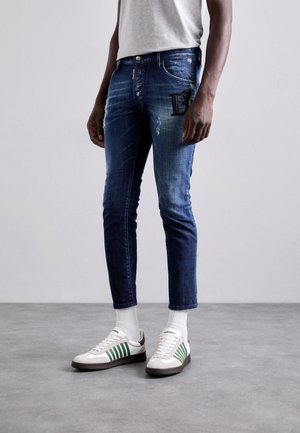 Jeans in denim blu, slim fit, con lievi graffi e dettagli con logo sulla tasca, abbinati a sneakers bianche con strisce verdi.