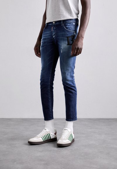 Blauwe denim jeans, slim fit, met lichte slijtage en logodetail op de zak, gecombineerd met witte sneakers met groene strepen.