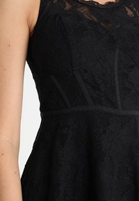 Robe noire sans manches avec motif en dentelle florale, corsage ajusté avec coutures verticales et détail de ceinture sur le haut du torse et le bras d’un mannequin.