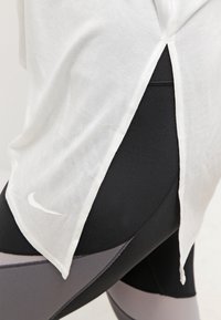 Witte, lichtgewicht T-shirt met zijsplitsingen gecombineerd met zwarte en grijze nauwsluitende leggings met een klein Nike-logo op het onderbeen.