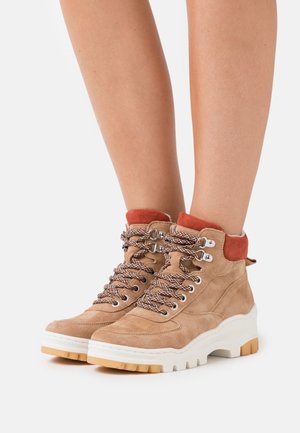 Braune Wildleder-Wanderschuhe mit weißer Gummisohle, gepolstertem roten Knöchelkragen und strukturierten braunen Schnürsenkeln. Mit silbernen Metallösen ausgestattet.