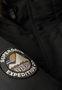 Tejido de chaqueta negra con un parche tejido que presenta montañas, un sol y el texto "Superdry Expedition Co." en marrón, blanco y beige.
