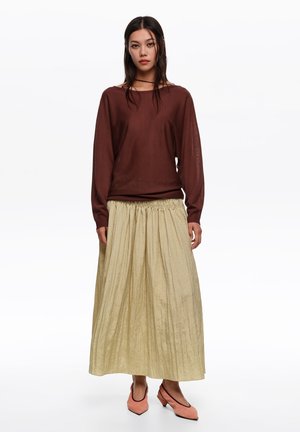 Kobieta ubrana w brązową bluzkę z długim rękawem, beżową plisowaną spódnicę midi i różowe buty na niskim obcasie, stojąca na białym tle.