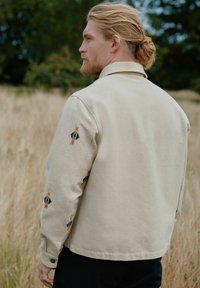 Beige puuvillajakku, jossa on mustat ja oranssit brodyyrit, jotka muodostavat geometrisiä kuvioita hihoissa. Takissa on klassinen kaulus ja nappikiinnitys.
