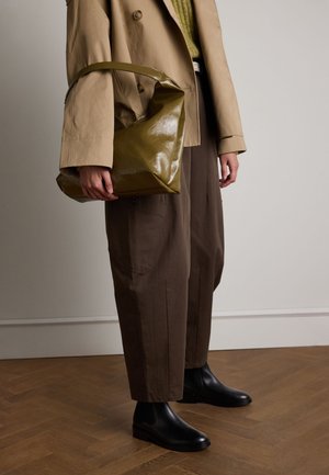 Sac en bandoulière vert olive et brillant avec une poignée courbée, porté avec un manteau beige à double boutonnage, un pantalon large marron et des bottines noires.