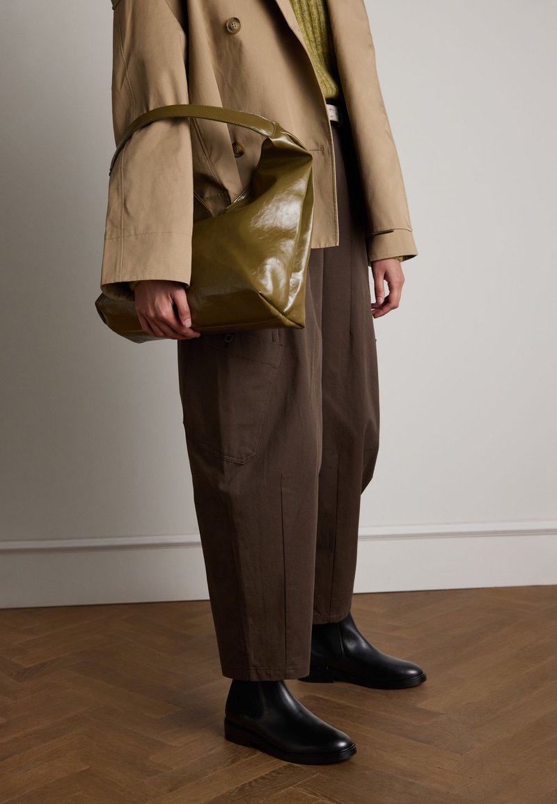 Borsa a spalla verde oliva, lucida, con manico curvo, indossata con un cappotto beige doppiopetto, pantaloni larghi marroni e stivali alla caviglia neri.