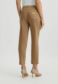 Femme portant un pantalon cropped beige et des sandales à talons hauts en cuir de serpent, se tenant devant un fond clair uni.