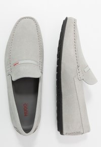 Mocassins en daim gris avec des détails cousus, un design à enfiler et une semelle en caoutchouc noire contrastante. Présente un petit logo rouge sur le côté.