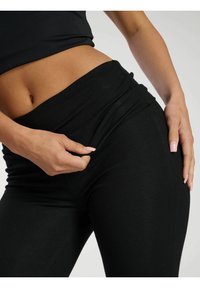 Schwarze gerippte Leggings mit hohem Bund, die eine glatte Textur aufweisen. Eine Hand passt den Stoff im Hüftbereich an.