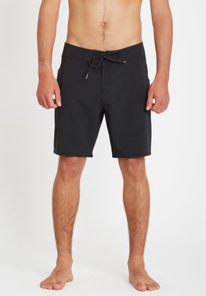 Modèle masculin portant un short de bain noir mi-cuisses avec taille à cordon, debout pieds nus sur un fond blanc uni.