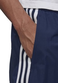 Námořnické šortky s bílými vertikálními pruhy, vybavené boční kapsou a elastickým pasem. Tkanina se zdá být lehká a hladká.