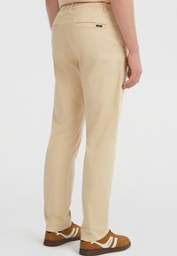 Persoon in beige rechte broek en bruine sneakers met witte strepen, staand tegen een effen lichte achtergrond.