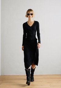 Haut noir côtelé avec fermeture éclair à l'avant, associé à une jupe portefeuille noire et des bottes noires à mi-mollet. La mannequin porte des lunettes de soleil. Fond en parquet.