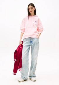 Sweat à capuche rose clair avec une poche avant et un logo brodé, associé à un jean large bleu clair et une veste bomber bordeaux.
