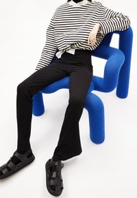 Černé žebrované kalhoty v kombinaci s pruhovaným dlouhým tričkem. Postava sedí na jasně modré sochařské židli, má na nohou černé sandály se pásky.