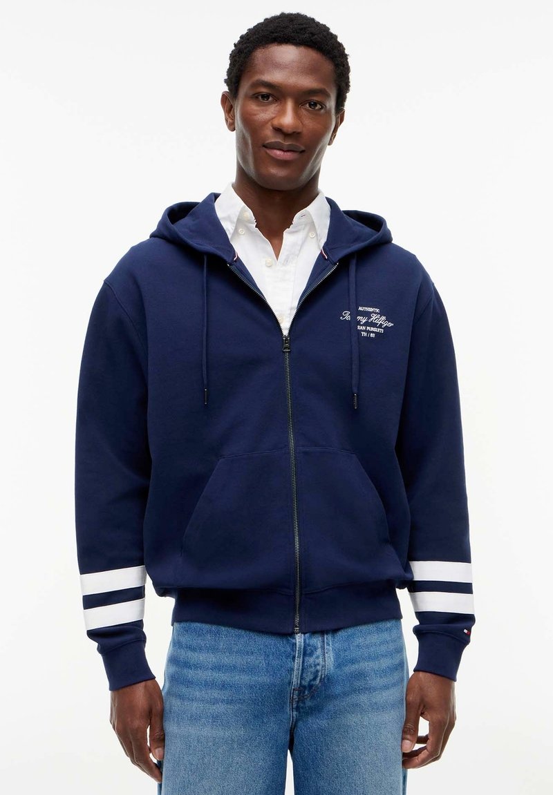 Homme portant un sweat à capuche bleu marine zippé avec des bandes blanches sur les manches et un jean bleu clair, debout devant un fond uni.