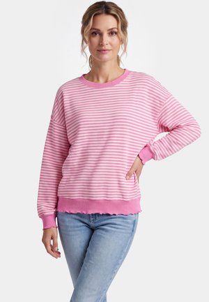 Frau steht mit der Hand an der Hüfte, trägt einen pink-weiß gestreiften Langarmpullover und hellblaue Jeans vor weißem Hintergrund.