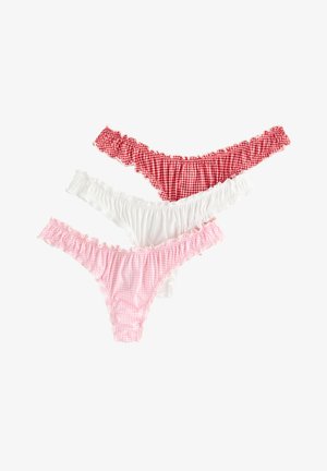 Jonge vrouw staat op blote voeten, draagt een wit geribd hemd en roze geruit ondergoed tegen een effen lichte achtergrond.