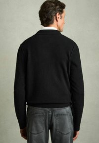 Zwarte wollen trui met een ontspannen pasvorm en een schipperkraag, gezien van achteren. Bevat lange mouwen en een gestructureerde afwerking. Grijze spijkerbroek zichtbaar daaronder.