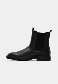 Botte Chelsea en cuir noir avec panneaux latéraux élastiques, languettes à l'avant et à l'arrière, et un petit talon sur fond gris clair.