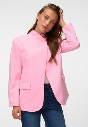 Femme aux longs cheveux bruns portant un blazer rose vif et un jean bleu, touchant ses cheveux avec une expression neutre.